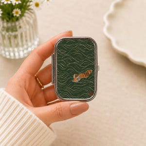 Könnte beinhalten: Eine rechteckige Pillendose aus Metall mit silbernem Rahmen und einem dunkelgrünen Deckel, der ein goldenes Wellenmuster sowie eine kleine orange-schwarze Tigerillustration in der Mitte des Designs aufweist.