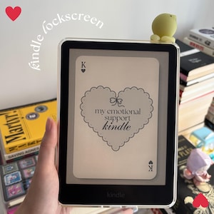 May include: A Kindle e-reader with a clear case, showing a custom lockscreen with a heart-shaped design and the text "my emotional support kindle." A small yellow chick figure sits on top. The words "kindle lockscreen" are written in cursive.