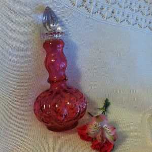 May include: A pink glass bottle with a clear glass stopper. The bottle has a textured surface and is shaped like a gourd. The bottle is sitting on a white crocheted surface with a pink flower.