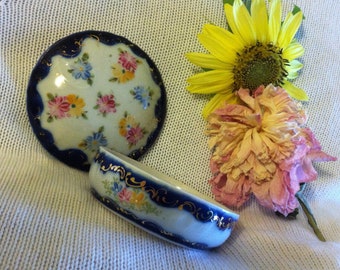 May include: A vintage porcelain trinket box with a blue and gold rim and floral designs. The lid is open and the box is sitting on a white knitted surface with dried flowers.