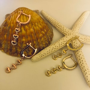 May include: A pair of gold hoop earrings with dangling charms. Each charm is a heart-shaped pendant with clear sparkling stones.