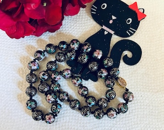 May include: A black necklace with round beads decorated with floral patterns in shades of pink, green, and gold. The beads are strung on a black cord.
