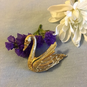 May include: A gold-tone swan brooch with detailed wings and a textured finish. The brooch is set against a light blue background with purple and white flowers.
