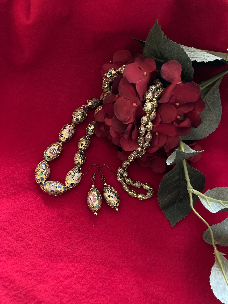 Puede incluir: Un collar y pendientes a juego de oro con dise&ntilde;os florales de esmalte de colores. El collar y los pendientes est&aacute;n sobre un fondo rojo con una flor roja.