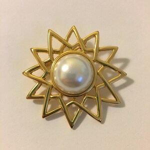 May include: A gold-toned brooch in the shape of a starburst, featuring a large, round, pearlescent white faux pearl at its center. The starburst design has pointed rays. The brooch is likely made of metal and is set against a white background.