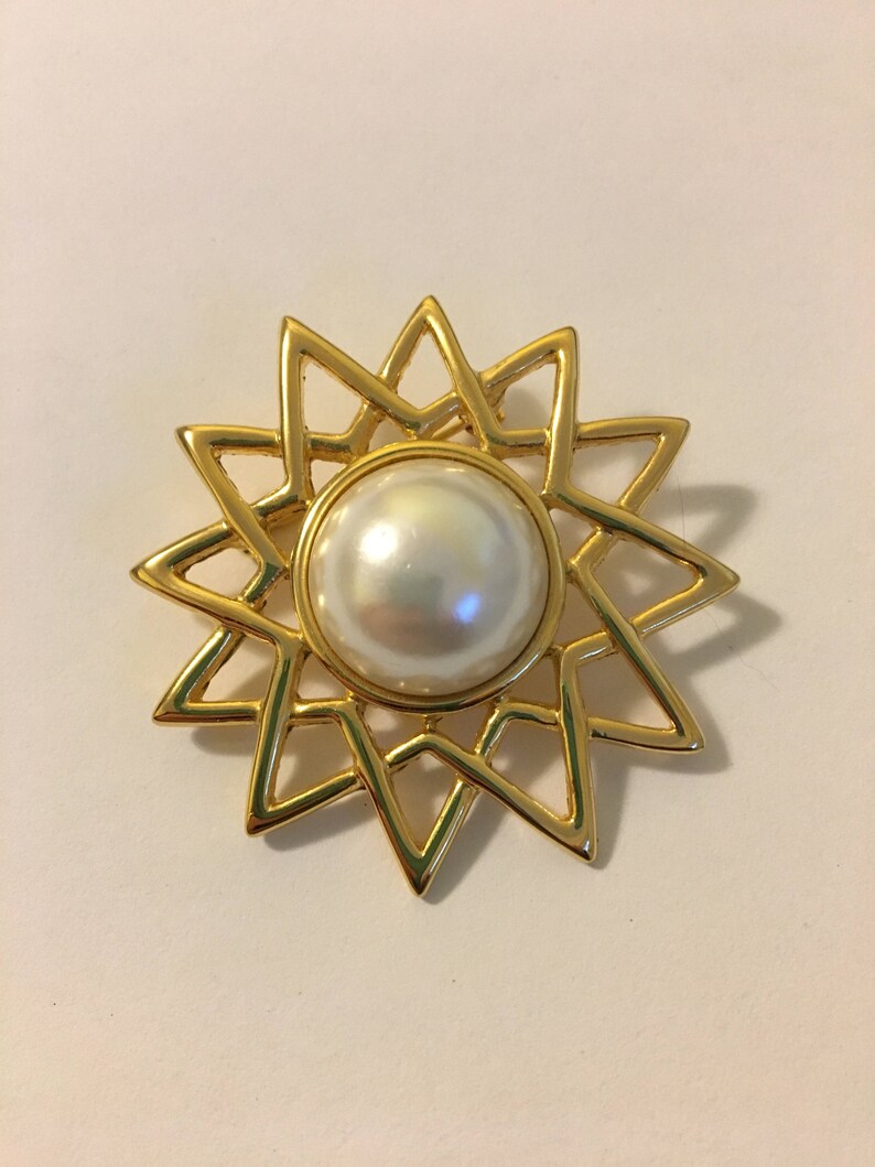 May include: A gold-toned brooch in the shape of a starburst, featuring a large, round, pearlescent white faux pearl at its center. The starburst design has pointed rays. The brooch is likely made of metal and is set against a white background.