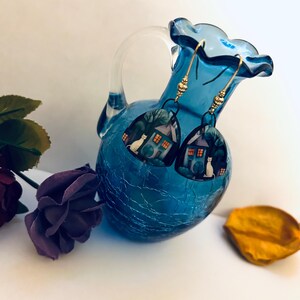 May include: A pair of dangle earrings featuring a house and cat design. The earrings are suspended from gold-colored hooks and are displayed near a blue glass vase and purple rose.