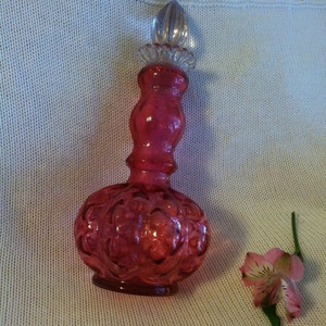 May include: A ruby red glass perfume bottle with a clear glass stopper. The bottle has a bulbous base with an embossed floral design. A pink and white flower with green stem rests beside the bottle.
