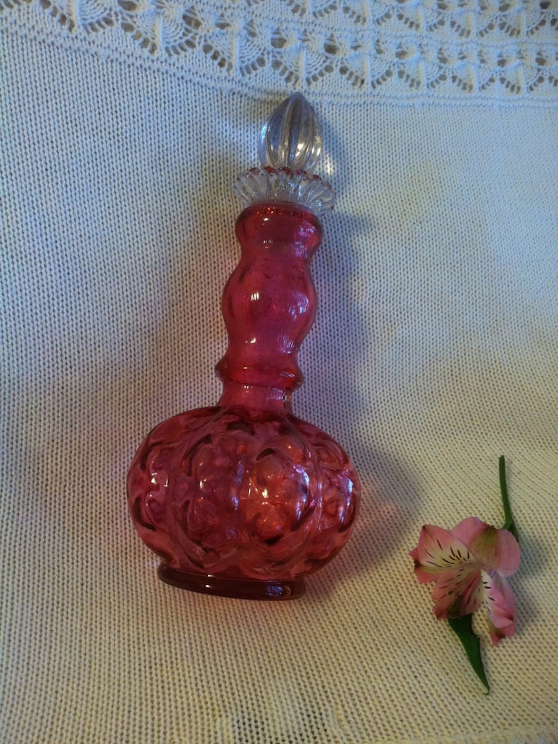May include: A ruby red glass perfume bottle with a clear glass stopper. The bottle has a bulbous base with an embossed floral design. A pink and white flower with green stem rests beside the bottle.