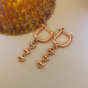 May include: A pair of rose gold earrings with a dangling chain of four heart-shaped charms. Each heart is accented with small clear stones.