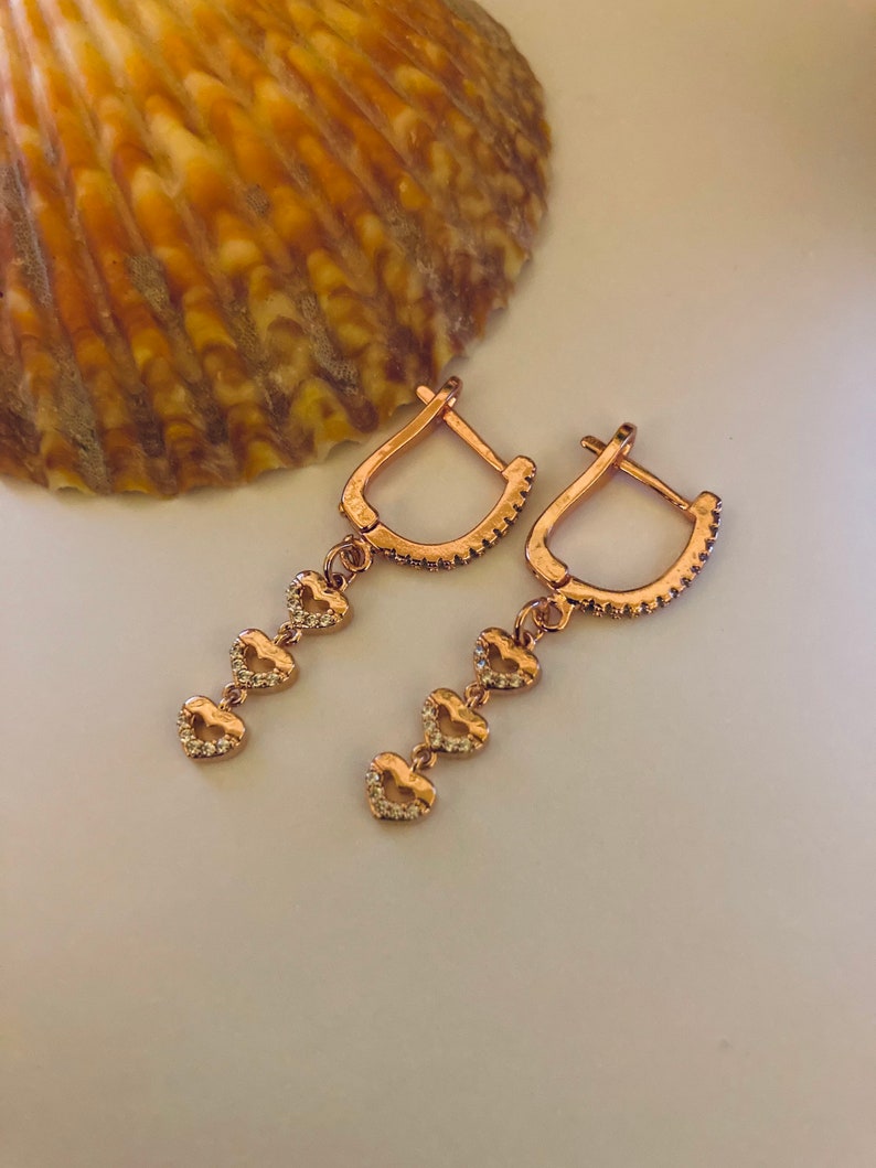 May include: A pair of rose gold earrings with a dangling chain of four heart-shaped charms. Each heart is accented with small clear stones.