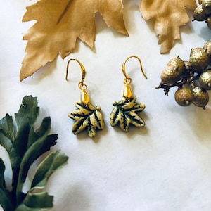 May include: A pair of gold-colored leaf earrings with green and gold detailing. The earrings are displayed on a white surface, with gold-colored leaves and berries in the background. The earrings have a hook closure.