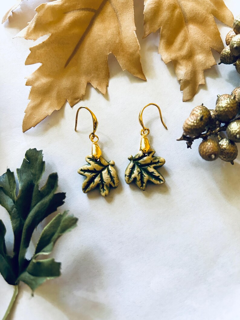 May include: A pair of gold-colored leaf earrings with green and gold detailing. The earrings are displayed on a white surface, with gold-colored leaves and berries in the background. The earrings have a hook closure.