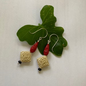 Puede incluir: Un par de pendientes colgantes con un dise&ntilde;o rojo y crema. Los pendientes presentan un dije de forma cuadrada con un dise&ntilde;o tallado y una cuenta en forma de l&aacute;grima.