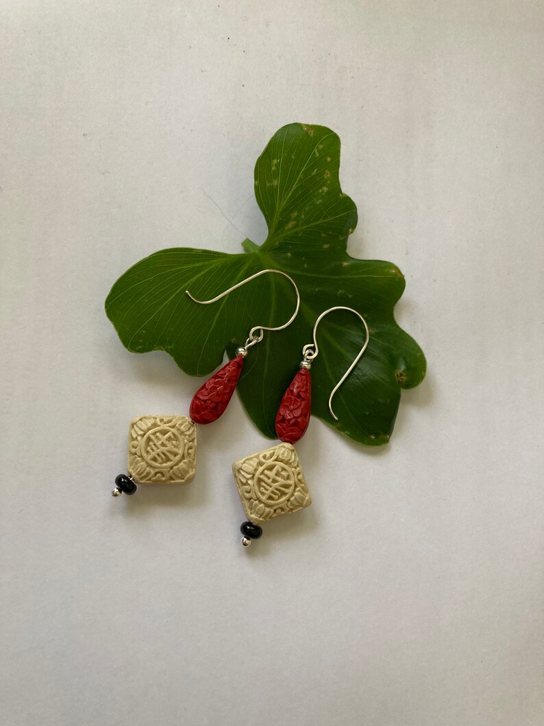 Puede incluir: Un par de pendientes colgantes con un dise&ntilde;o rojo y crema. Los pendientes presentan un dije de forma cuadrada con un dise&ntilde;o tallado y una cuenta en forma de l&aacute;grima.