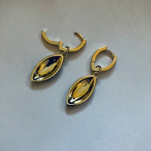 May include: A pair of gold hoop earrings with a teardrop-shaped pendant. The pendants are a dark gold color and have a smooth, shiny finish.