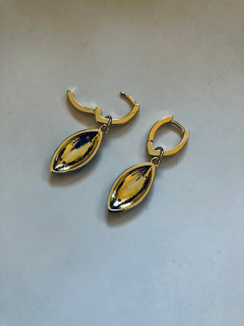 May include: A pair of gold hoop earrings with a teardrop-shaped pendant. The pendants are a dark gold color and have a smooth, shiny finish.