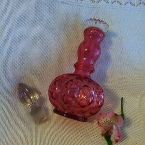 May include: A vintage, ruby-red glass perfume bottle with a clear glass stopper. The bottle has a bulbous base and a long, curved neck, with a decorative floral pattern. A pink and white flower rests beside the bottle.