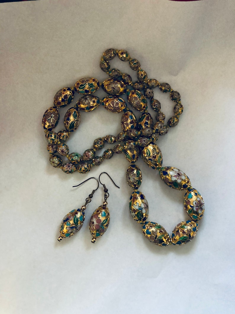 Puede incluir: Un collar y pendientes de oro con cuentas de forma ovalada. Las cuentas est&aacute;n decoradas con motivos florales en tonos rosa, azul, verde y blanco.