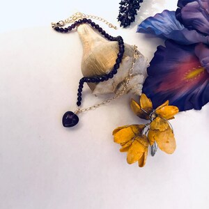 May include: A delicate gold chain necklace with a dark purple heart pendant, accented with a strand of dark purple beads. The necklace is displayed on a seashell with purple and yellow flowers in the background.