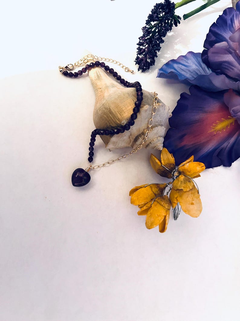 May include: A delicate gold chain necklace with a dark purple heart pendant, accented with a strand of dark purple beads. The necklace is displayed on a seashell with purple and yellow flowers in the background.