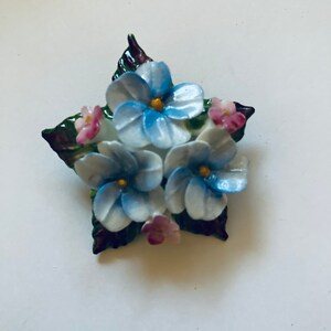 May include: A porcelain brooch featuring a cluster of blue and white flowers with pink accents. The flowers are surrounded by green leaves.