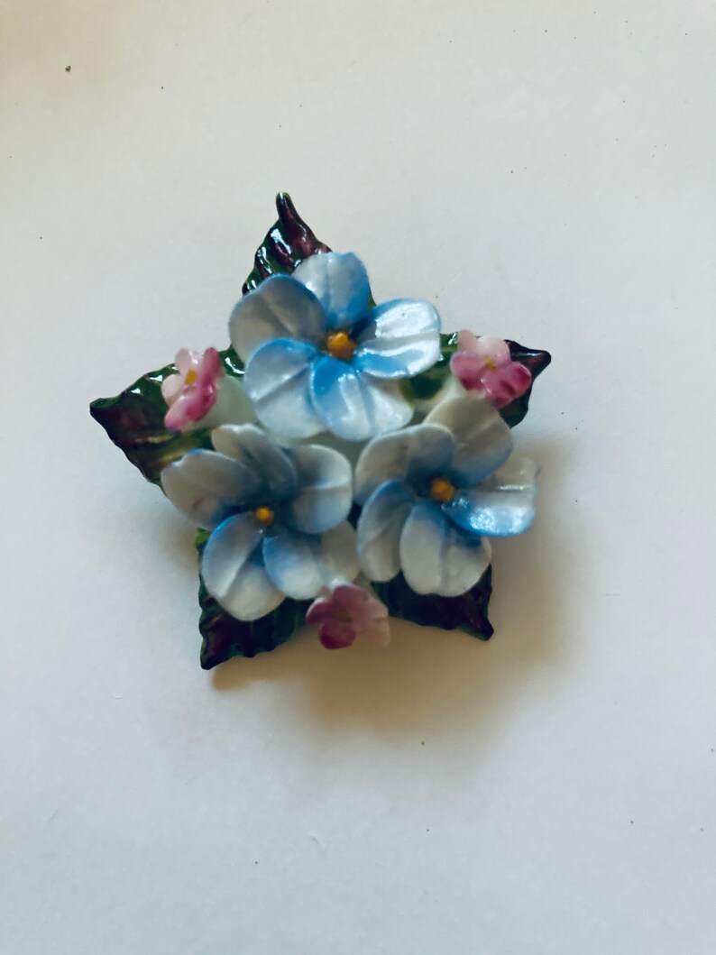 May include: A porcelain brooch featuring a cluster of blue and white flowers with pink accents. The flowers are surrounded by green leaves.