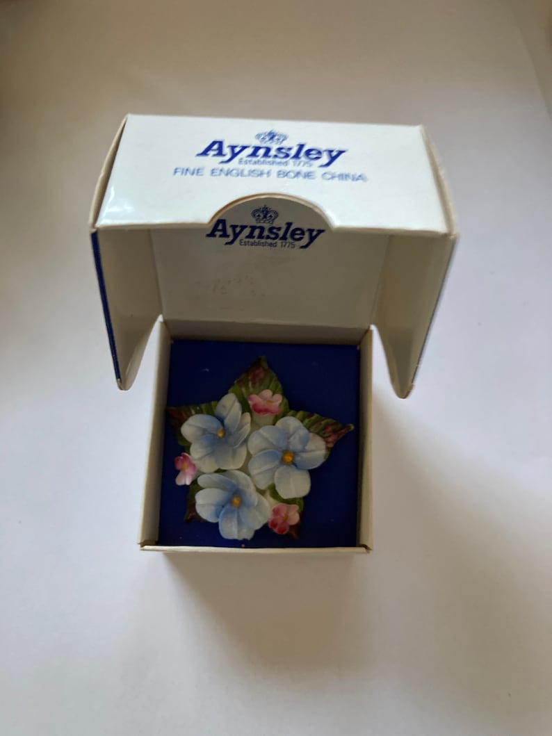 May include: A blue and white porcelain flower brooch in a star shape, in a white box with the words "Aysnley Established 1775 Fine English Bone China" on the lid.