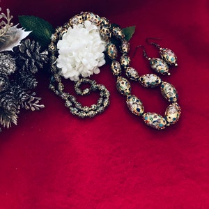 Puede incluir: Un collar y pendientes de oro con cuentas ovaladas decoradas con motivos florales coloridos. Las cuentas est&aacute;n colocadas sobre un fondo rojo.