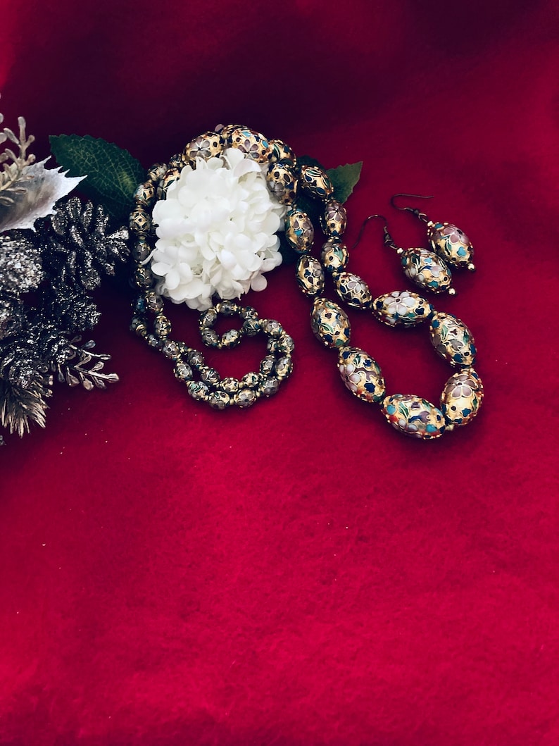 Puede incluir: Un collar y pendientes de oro con cuentas ovaladas decoradas con motivos florales coloridos. Las cuentas est&aacute;n colocadas sobre un fondo rojo.