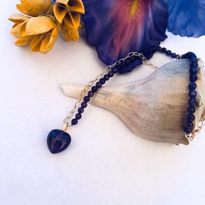 May include: A gold chain necklace with a purple heart pendant and purple beads. The necklace is displayed on a seashell with yellow flowers in the background.