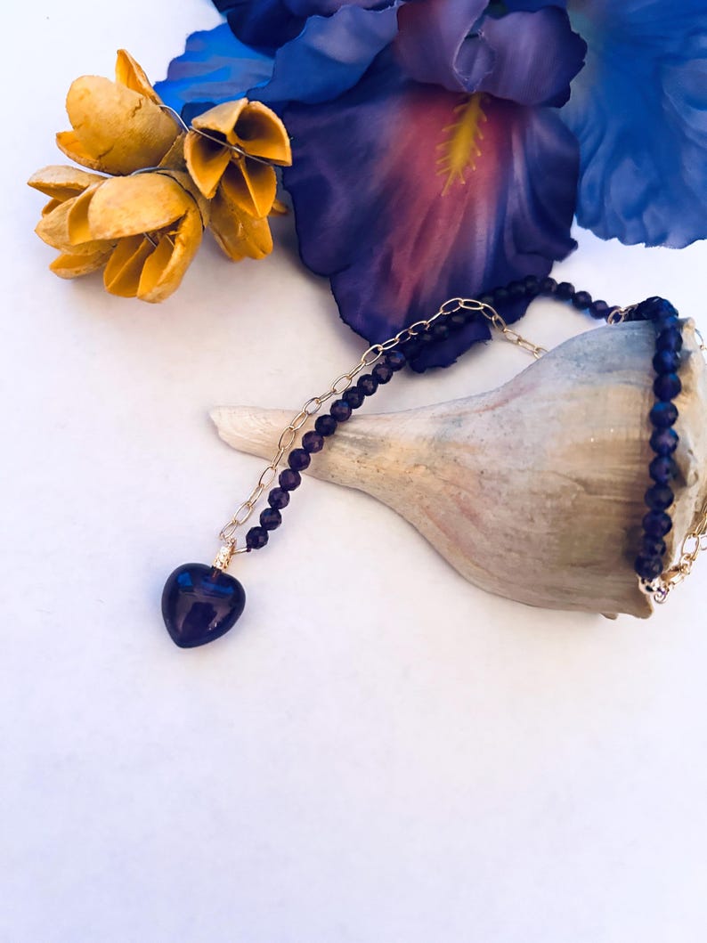 May include: A gold chain necklace with a purple heart pendant and purple beads. The necklace is displayed on a seashell with yellow flowers in the background.