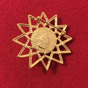 May include: A gold-toned starburst brooch with a textured center and openwork design. The back features a pin clasp for secure attachment. The brooch is set against a red fabric background.