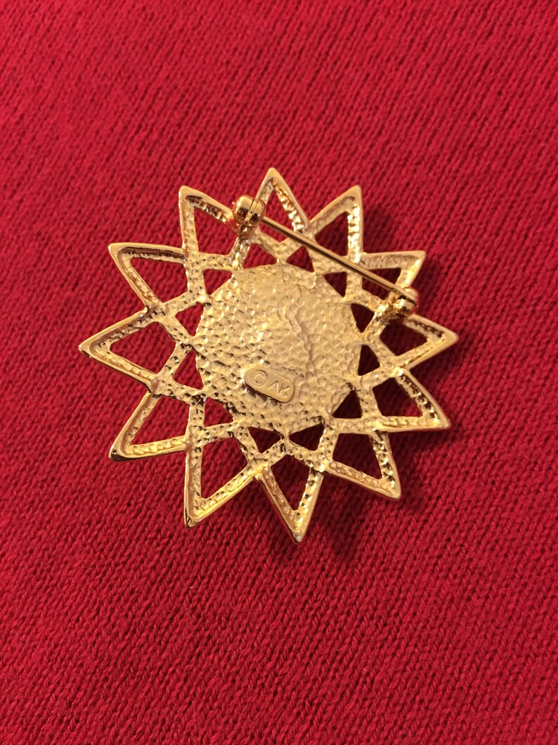 May include: A gold-toned starburst brooch with a textured center and openwork design. The back features a pin clasp for secure attachment. The brooch is set against a red fabric background.