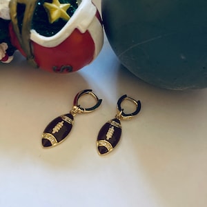 May include: A pair of gold hoop earrings with a brown football charm. The football charm has white stitching and a row of clear rhinestones around the edge.