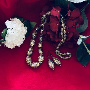 Puede incluir: Un conjunto de pendientes y collar de tono dorado con cuentas de forma ovalada. Las cuentas est&aacute;n decoradas con motivos florales en tonos azules, rosas y blancos. La joyer&iacute;a se muestra sobre un fondo de terciopelo rojo.