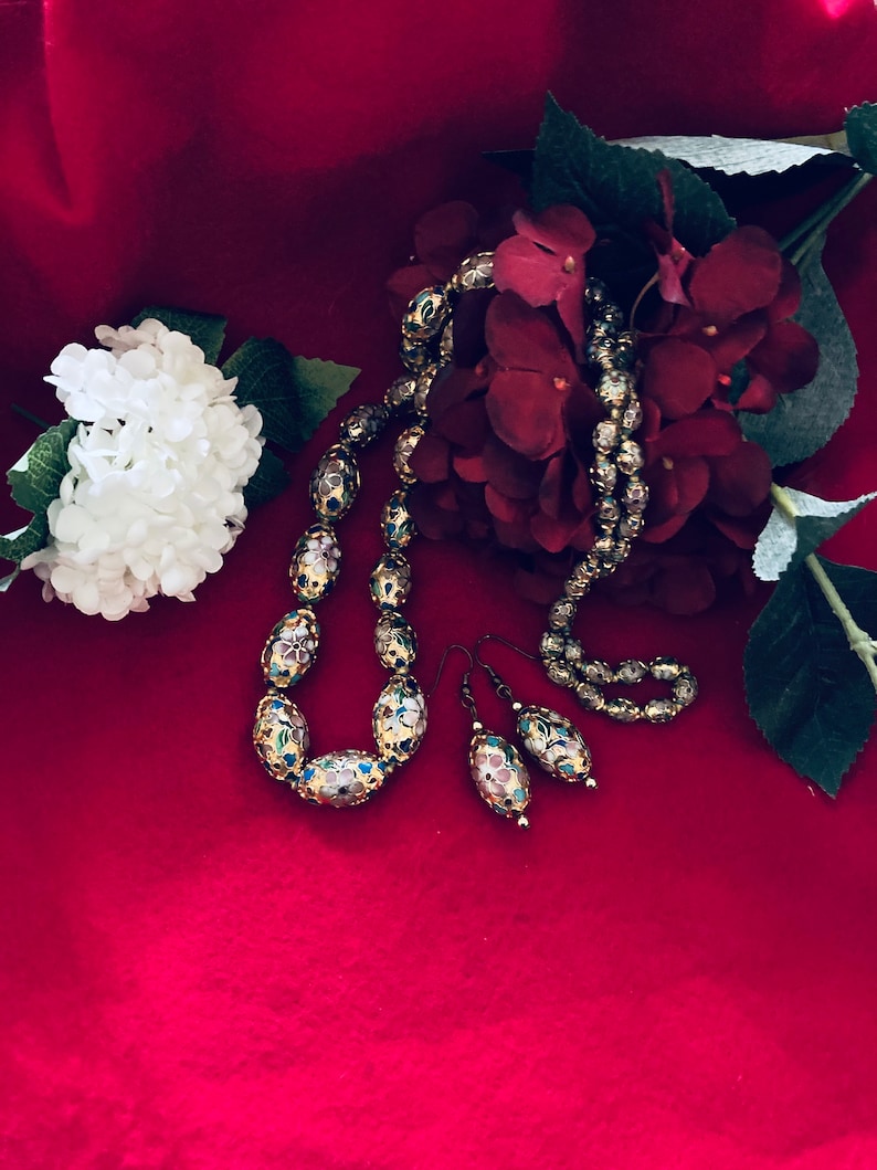 Puede incluir: Un conjunto de pendientes y collar de tono dorado con cuentas de forma ovalada. Las cuentas est&aacute;n decoradas con motivos florales en tonos azules, rosas y blancos. La joyer&iacute;a se muestra sobre un fondo de terciopelo rojo.
