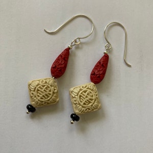 Puede incluir: Un par de pendientes de plata con cuentas talladas en rojo y blanco. Las cuentas blancas tienen forma cuadrada y presentan un car&aacute;cter chino. Los pendientes cuelgan de ganchos de plata.