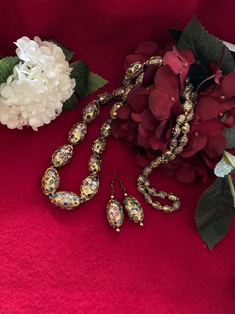 Puede incluir: Un collar y pendientes de oro con cuentas ovaladas decoradas con motivos florales de esmalte de colores. El collar y los pendientes est&aacute;n sobre un fondo rojo con flores blancas y rojas.