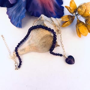 May include: A purple amethyst heart pendant necklace with a gold chain and purple amethyst beads. The necklace is displayed on a seashell.