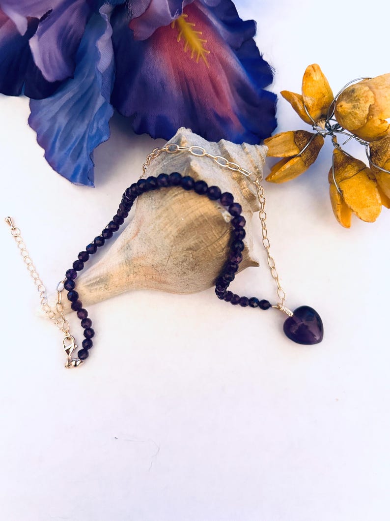 May include: A purple amethyst heart pendant necklace with a gold chain and purple amethyst beads. The necklace is displayed on a seashell.