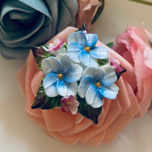 May include: A porcelain brooch featuring a cluster of blue and white flowers with green leaves and pink accents. The flowers are arranged on a pink rose.