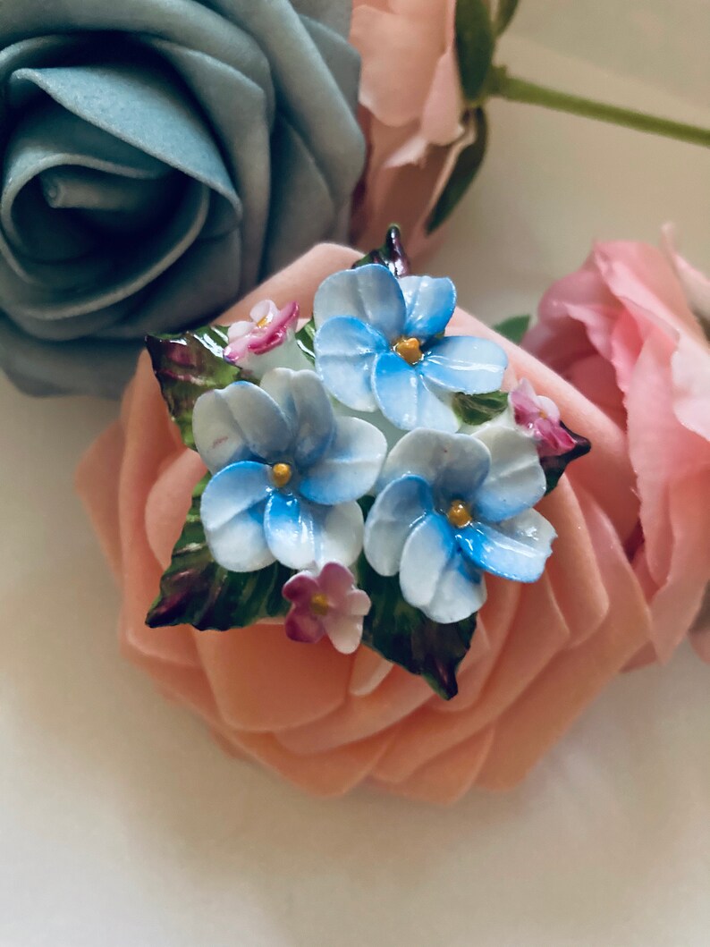 May include: A porcelain brooch featuring a cluster of blue and white flowers with green leaves and pink accents. The flowers are arranged on a pink rose.