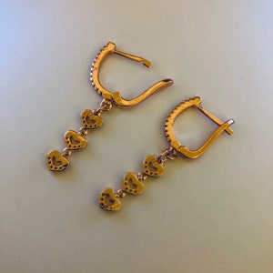 May include: A pair of gold-tone dangle earrings with a heart-shaped design. Each earring features four small hearts hanging from a hoop.