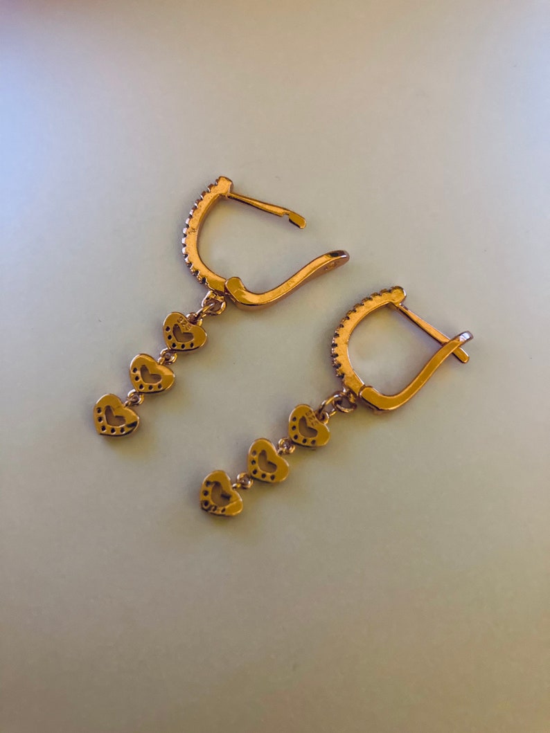 May include: A pair of gold-tone dangle earrings with a heart-shaped design. Each earring features four small hearts hanging from a hoop.