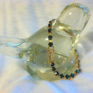 May include: A gold-toned bracelet with emerald-green square-cut stones. The bracelet is displayed on a clear glass sculpture. The clasp is visible, and the bracelet appears to be new.