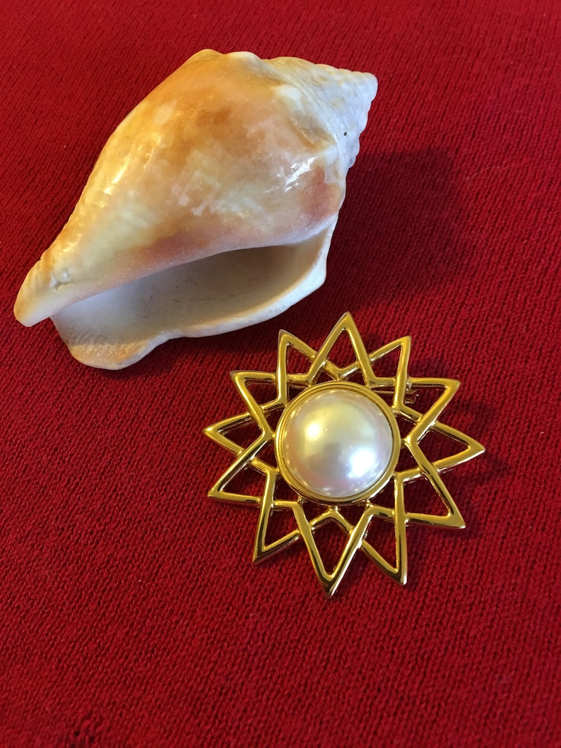 May include: A gold-toned starburst brooch with a large, round, white pearl center. A large, cream-colored seashell is positioned above the brooch on a red background. The brooch has a geometric design.