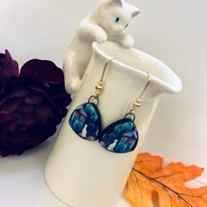 May include: A pair of dangle earrings with a whimsical design. Each earring features a dark blue, teardrop-shaped pendant with a white cat in front of a house. The earrings are suspended from gold-colored hooks.