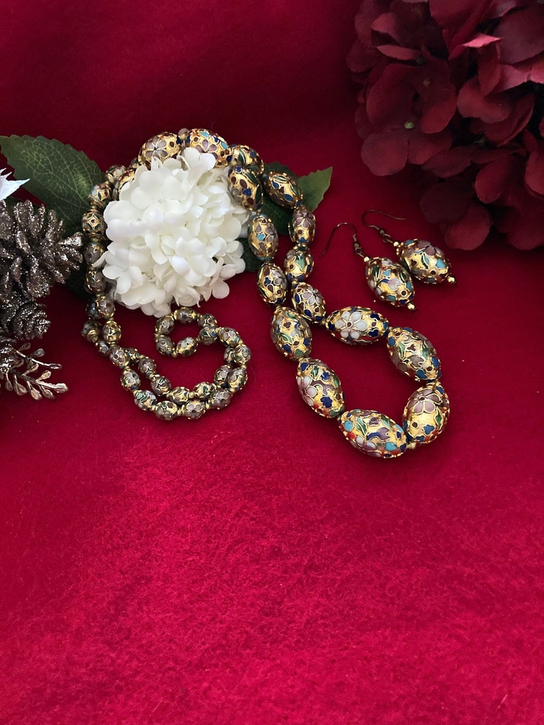 Puede incluir: Un collar y pendientes de oro con cuentas ovaladas decoradas con motivos florales de esmalte de colores. Las cuentas est&aacute;n sobre un fondo de terciopelo rojo.