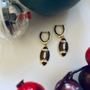May include: A pair of gold hoop earrings with a brown football charm. The football charm has white stitching and a small diamond accent.